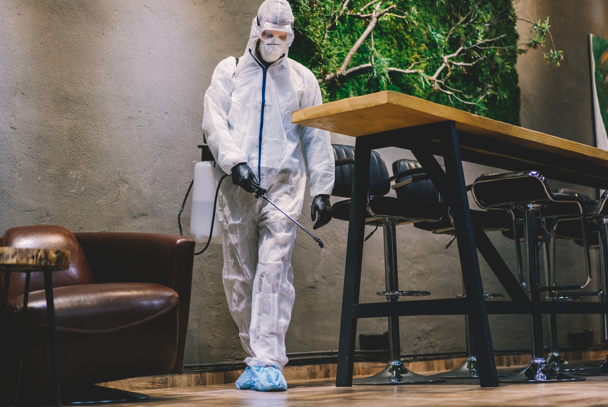 Pest control technician wearing a white protective suit, mask and gloves spraying pest control treatment along the floor of a commercial indoor space.